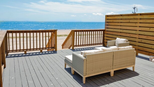 A wooden deck with a wicker sofa and a small table, overlooking a beach with a clear blue sky.