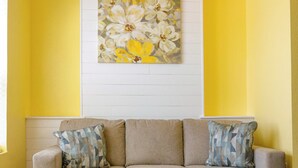 A beige sofa with two patterned cushions against a yellow wall, a floral painting above, and a wooden floor.