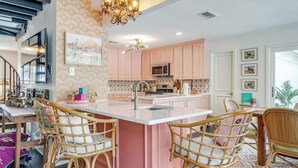 A modern kitchen with pink cabinets, a central island, and a dining area with wicker chairs. - Private Pool Oasis | Spa & Game Room • 12 (Fort Worth)