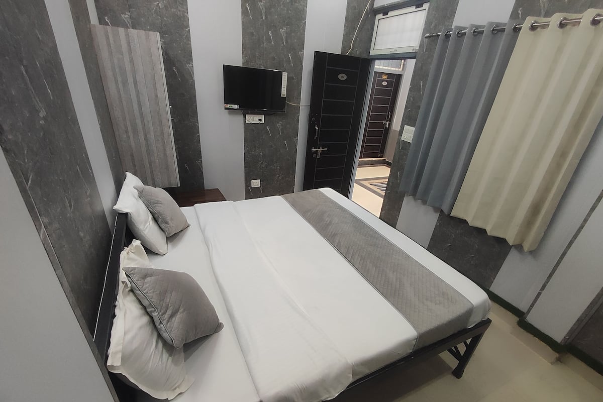 A single-bed room with a flat-screen TV mounted on the wall, a door with a number plate, and a curtain on the window.