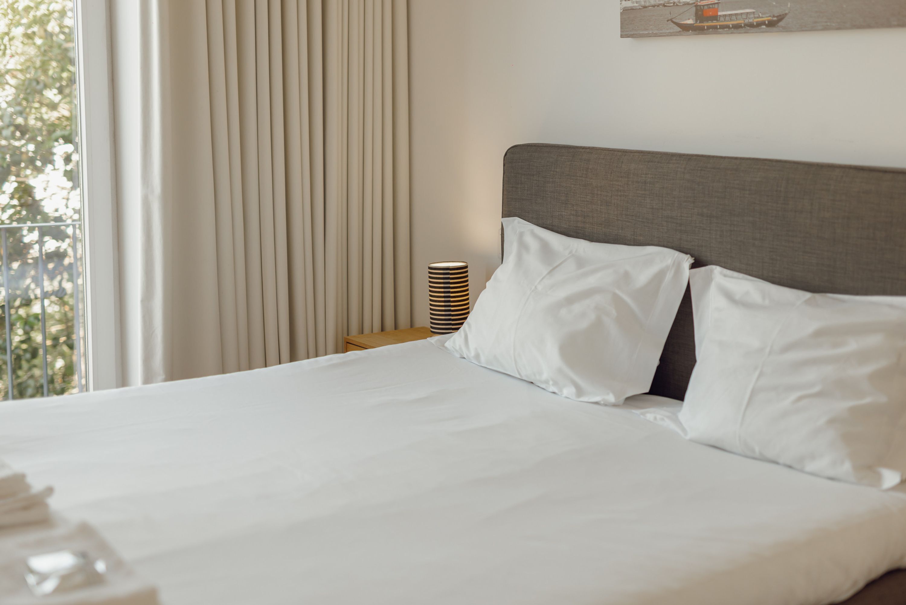 Un lit bien fait, avec du linge de lit blanc, une tête de lit grise et une table de chevet sur laquelle se trouve une lampe à rayures.