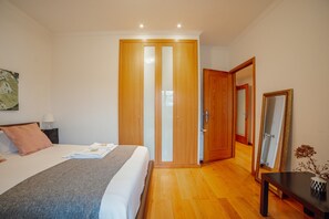Un dormitorio con cama, un armario de madera, una mesita de noche, una mesa con un florero y un espejo.