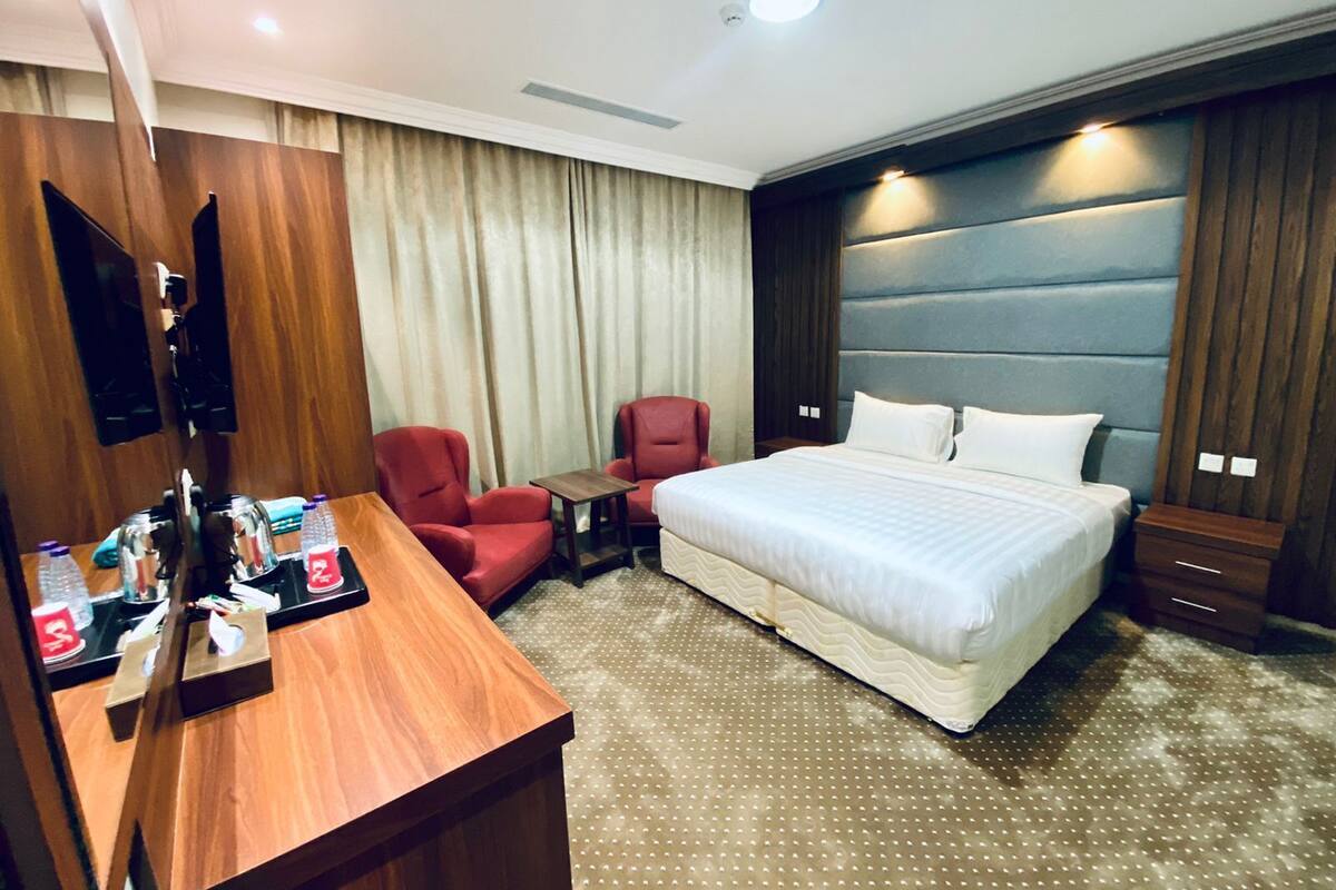 Habitación de hotel con una cama grande, dos sillones rojos, una mesita pequeña, un televisor y un escritorio de madera con detalles.