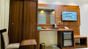 A hotel room with a wooden wardrobe, a desk with a computer, a TV mounted on the wall, and a chair.