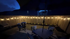 Ein Terrassenbereich im Freien mit Holzzaun, Lichterkette, Grill und Sitzgelegenheiten.
