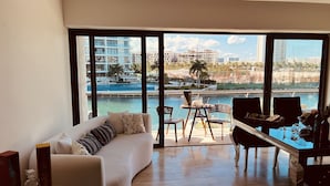 Un salon moderne avec un canapé blanc, une table basse en verre et un coin repas avec une table dressée pour deux personnes. De grandes fenêtres offrent une vue sur une piscine et les bâtiments environnants.