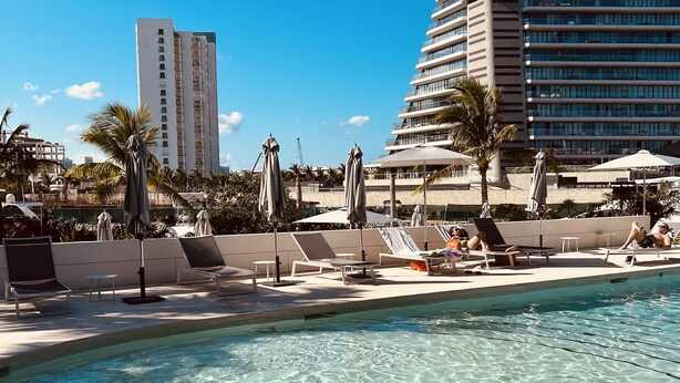 Un espace piscine doté de chaises longues et de parasols, entouré de palmiers et de bâtiments modernes.