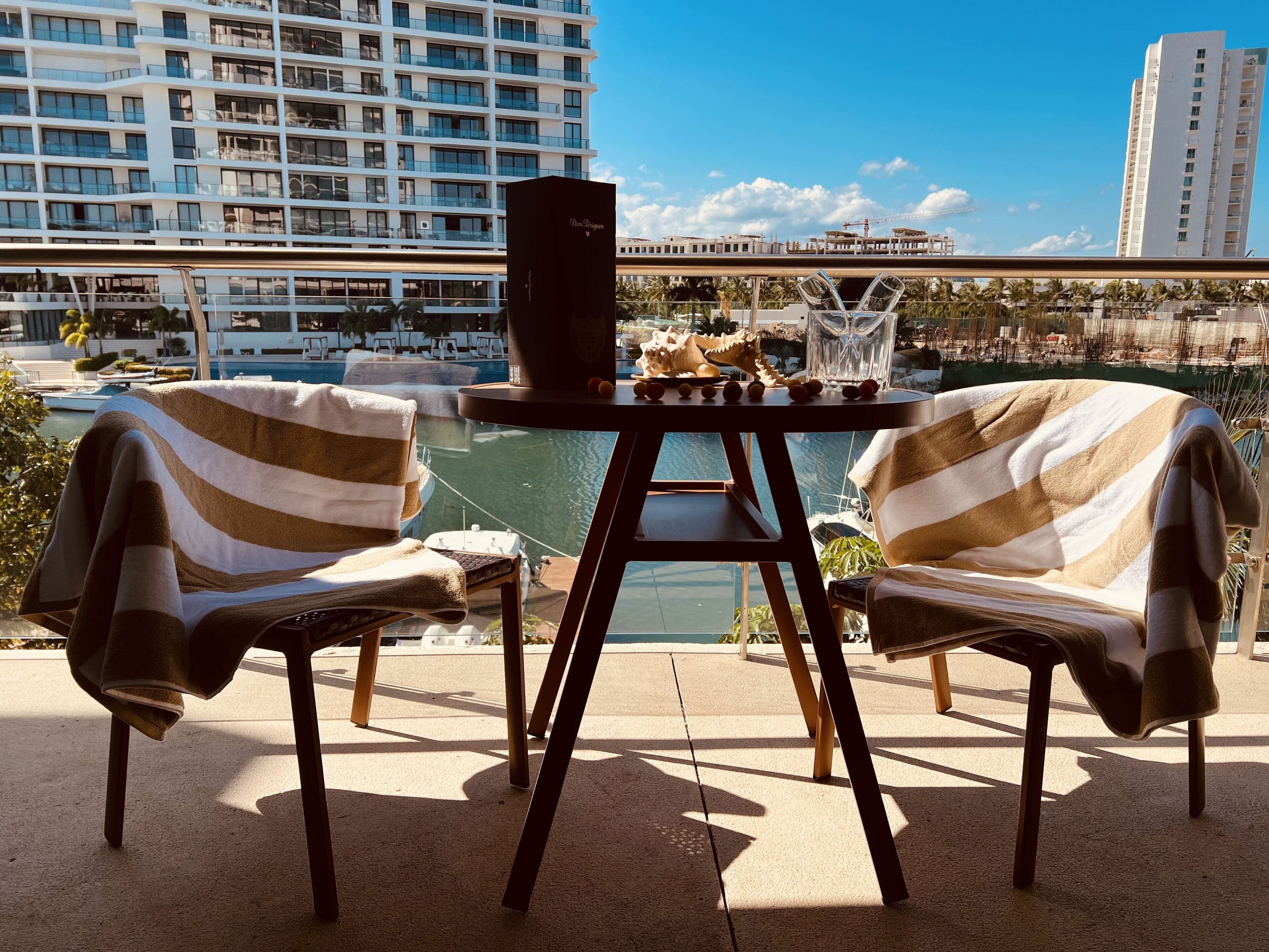A terrace with two striped lounge chairs, a table with a speaker and decorative items, overlooking a marina and buildings.