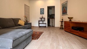 Un salon moderne avec un canapé gris, une petite table d’appoint sur laquelle est posé un vase, un meuble en bois et un coin cuisine visible à travers une porte ouverte.