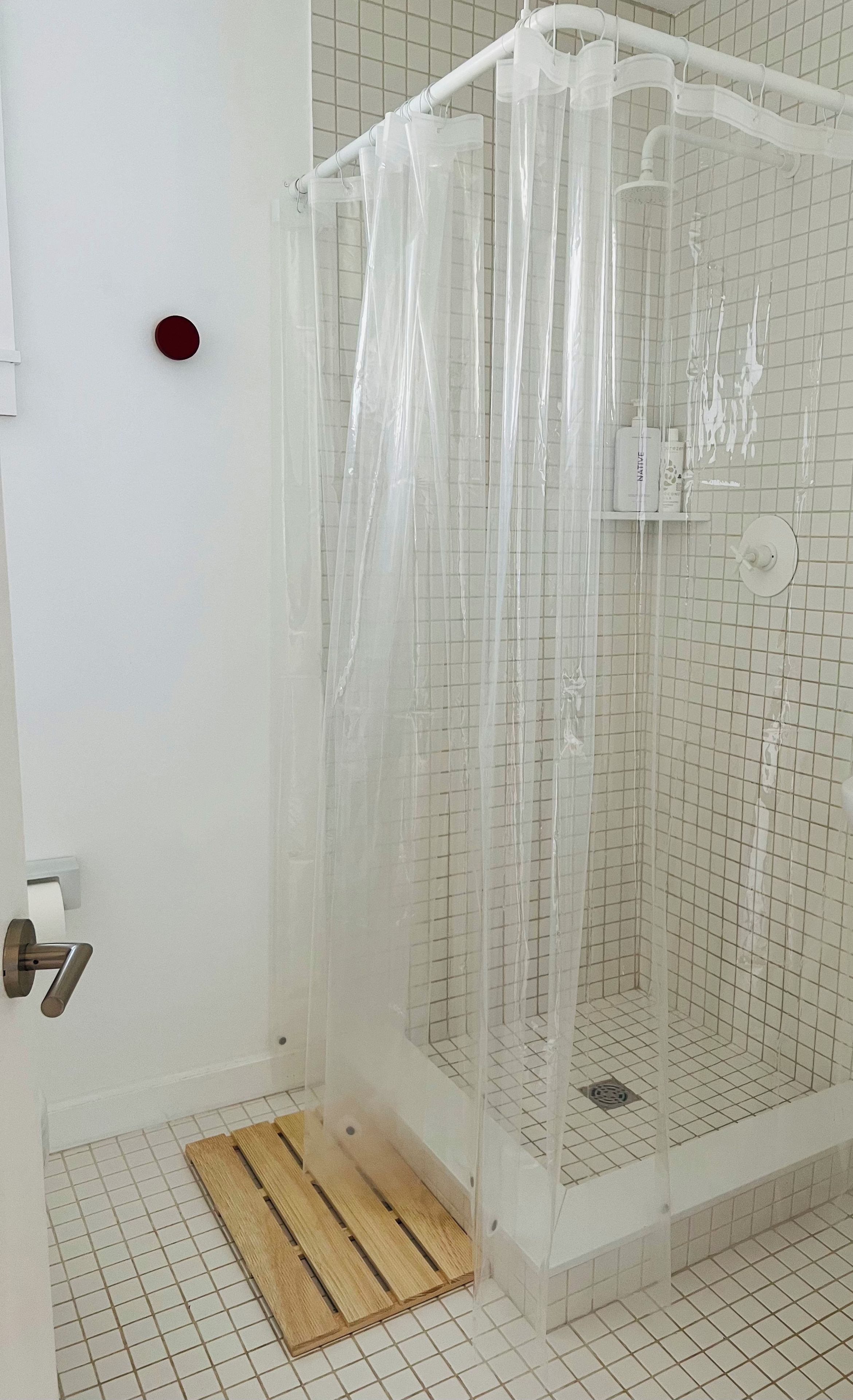 Un espace douche doté d’un rideau transparent, d’un tapis en bois à l’entrée et d’un sol carrelé à motifs géométriques.