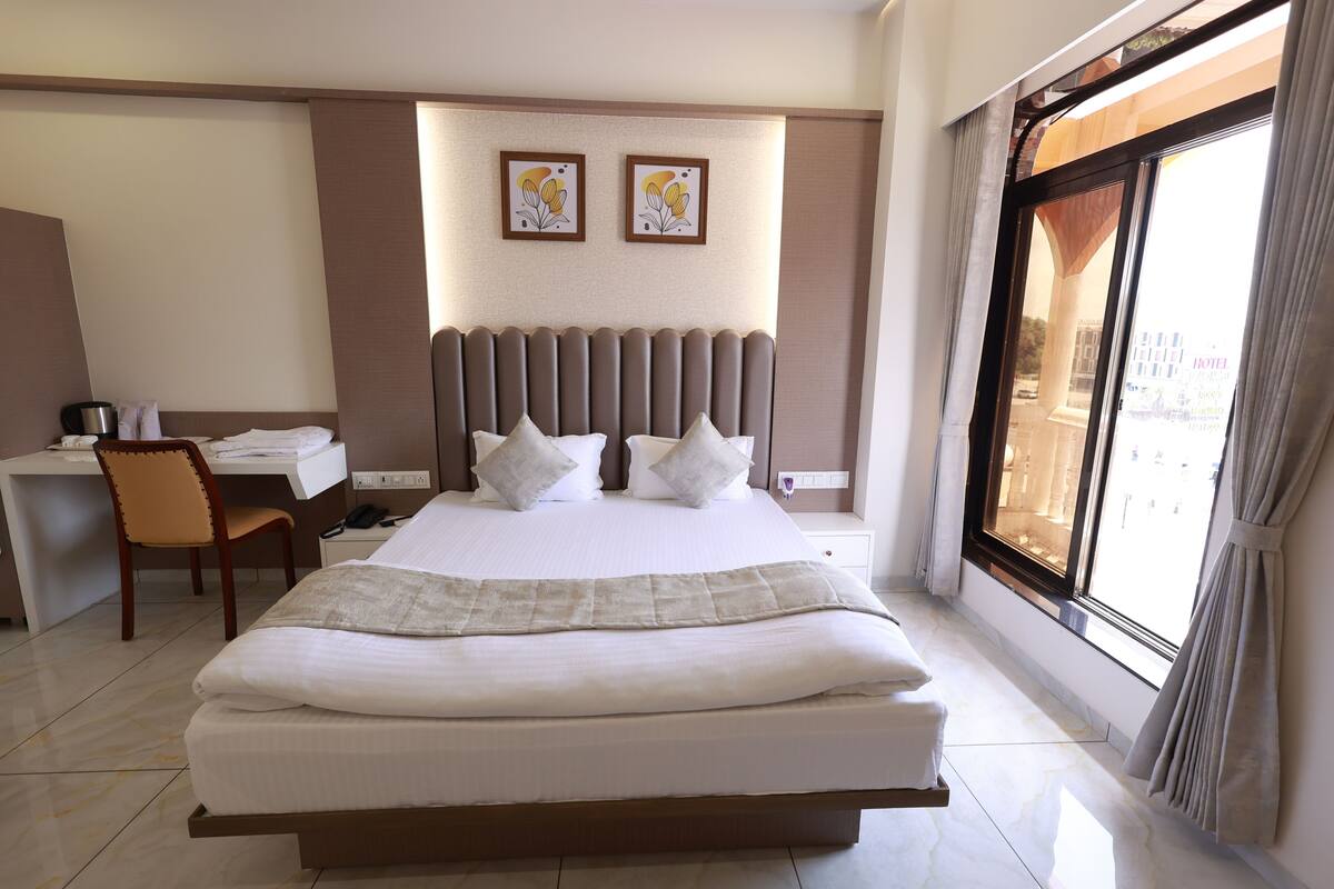 A hotel room with a large bed, a desk, a chair, and a view of the city through a large window.