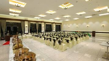 Un salón de banquetes con filas de sillas cubiertas de blanco, una larga mesa de buffet con vajilla dorada y candelabros en el techo.