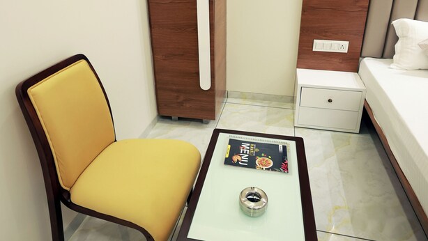 A small room with a yellow chair, a white nightstand, a white drawer, a wooden wardrobe, and a glass-top coffee table with a magazine. - HOTEL GRAND HAVELI (Umargam)