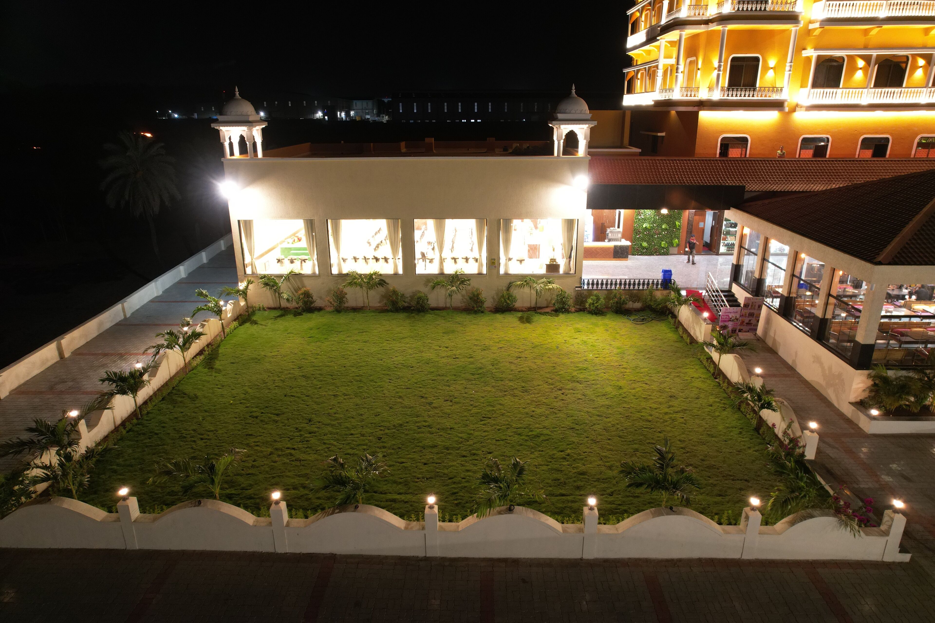 A hotel courtyard at night with a well-lit lawn, a white building with multiple windows, and a balcony overlooking the area.
