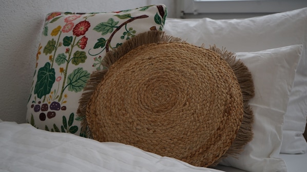 A bed with white bedding, a woven round cushion, and a floral pillow.