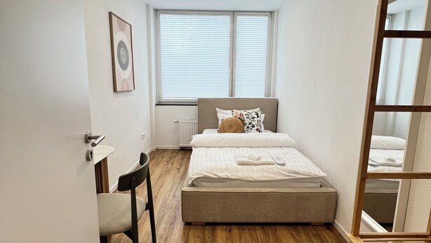 A bedroom with a bed, a chair, a desk, and a window with blinds. - House of Al-Ay Künzelsau (Künzelsau)