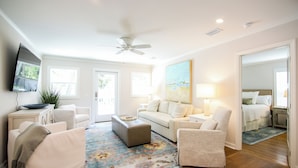 A spacious living room with a ceiling fan, a large painting, and a view into the bedroom through an open door.