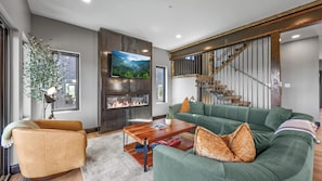 A modern living room with a green sofa, a fireplace, a TV, and a staircase.
