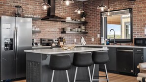A modern kitchen with a brick wall, stainless steel appliances, and a large island with bar stools. - The Back Nine Retreat by Avantstay Private Golf Sim, Firepit & Mountain Views Near Dollywood (Sevierville)