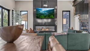 A modern living room with a large flat-screen TV mounted on a wall above a fireplace, a green sofa with cushions, and a large window providing a view of the outdoors. - The Back Nine Retreat by Avantstay Private Golf Sim, Firepit & Mountain Views Near Dollywood (Sevierville)