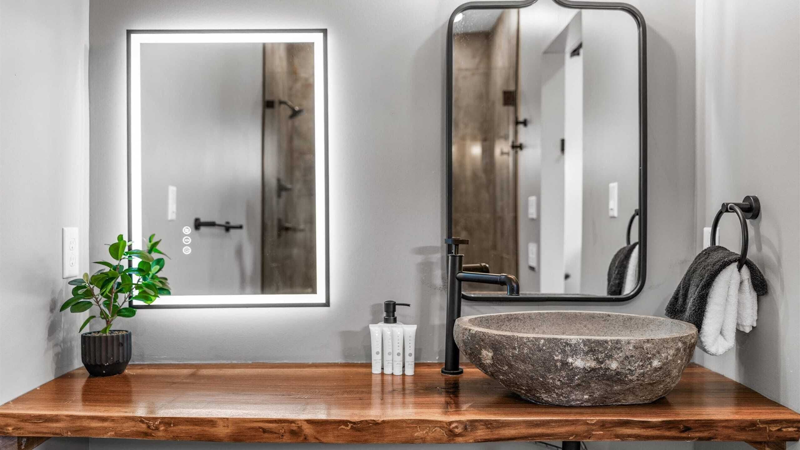 Un baño con tocador de madera, lavamanos de piedra, espejo, planta en maceta y toallero.