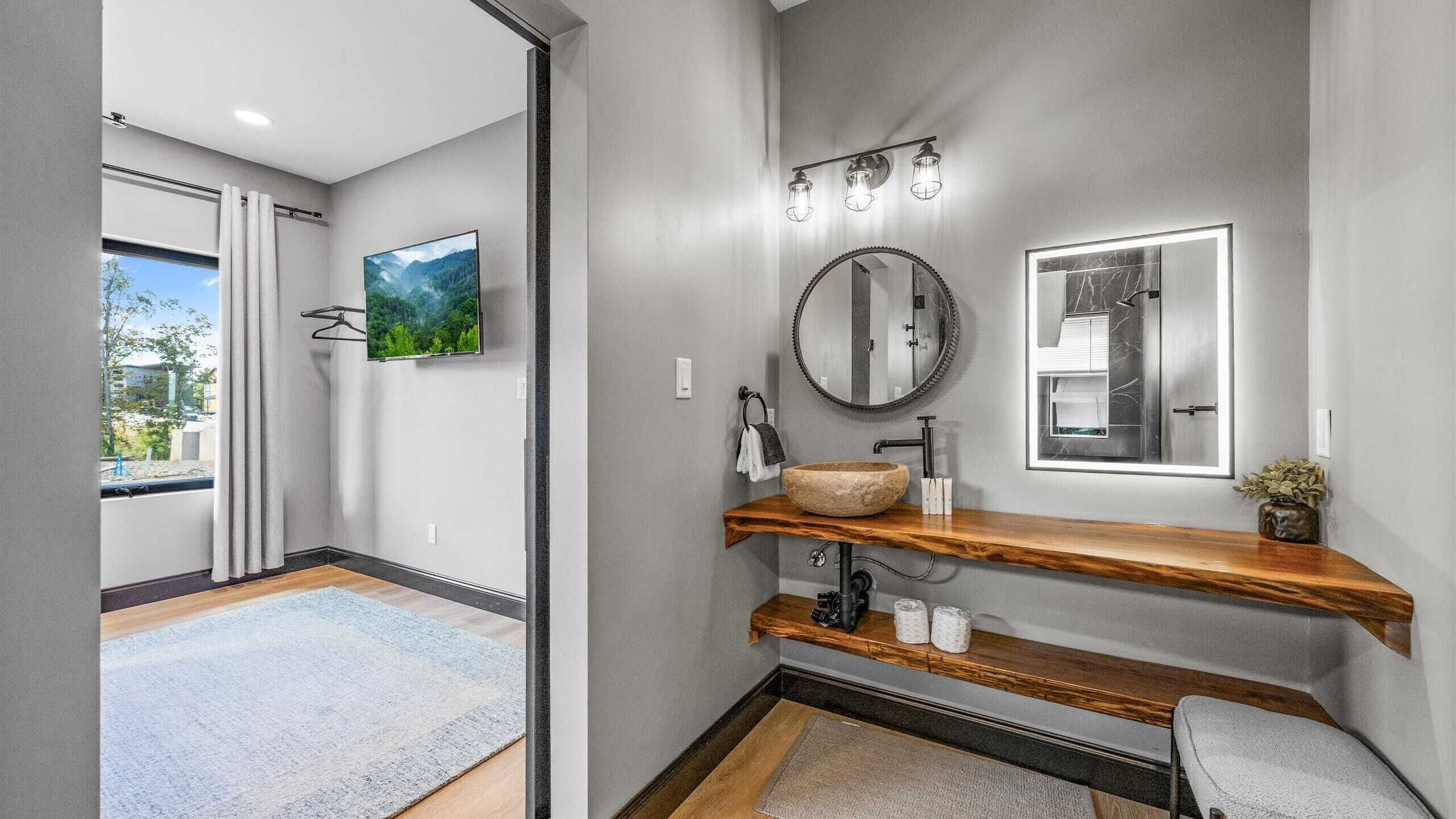 Une salle de bain moderne avec un meuble-lavabo en bois, un miroir rond et une grande fenêtre donnant sur un paysage verdoyant.