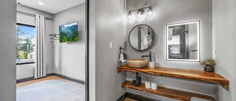 Une salle de bain moderne avec un meuble-lavabo en bois, un miroir rond et une grande fenêtre donnant sur un paysage verdoyant.