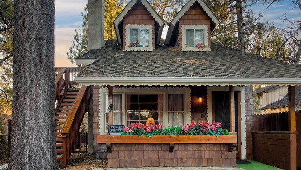 A small, two-story wooden cabin with a porch, a staircase, and a stone foundation.