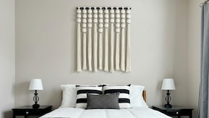 A modern bedroom with a bed, black and white striped pillows, a gray throw, and a macrame wall hanging.