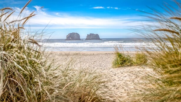 A beach with two large rock formations in the distance, a sandy foreground, and a clear blue sky.