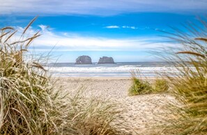 A beach with two large rock formations in the distance, a sandy foreground, and a clear blue sky.