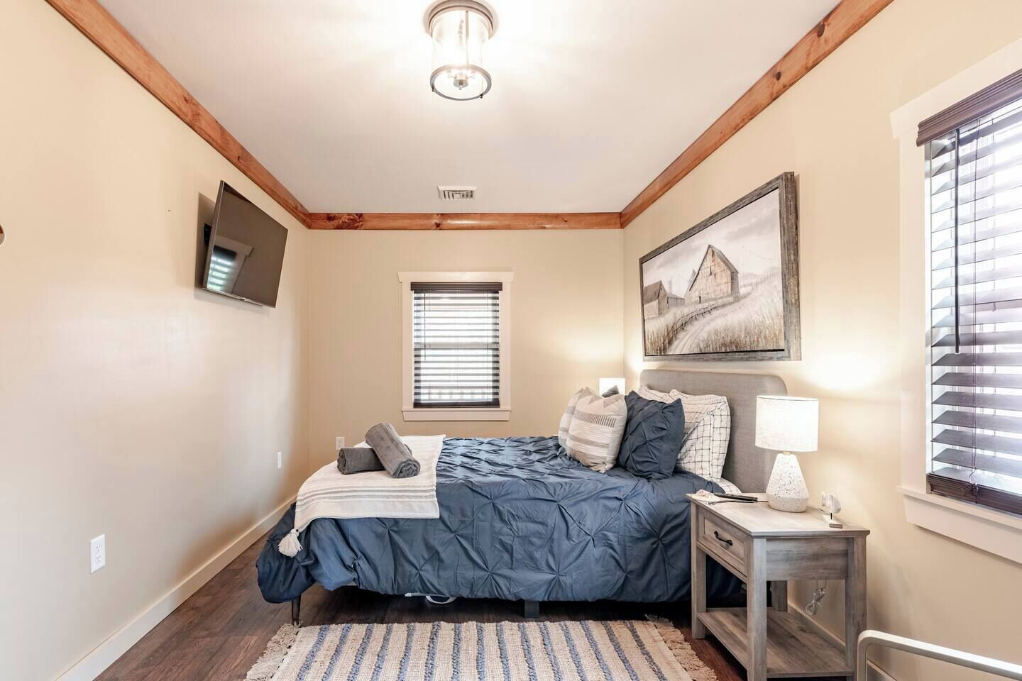 A bedroom with a bed, a nightstand, a lamp, a painting, a window with blinds, and a ceiling light.