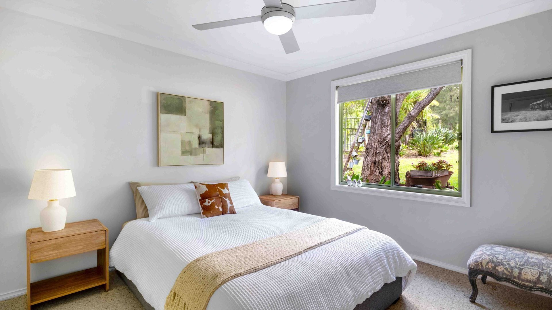 Una camera da letto con un letto, due comodini con lampade, una ventola a soffitto, una grande finestra con vista sull'esterno e un quadro appeso al muro.