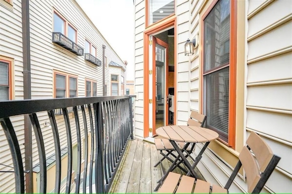 Un balcón con mesa y sillas, barandilla y un edificio con ventanas y puerta.