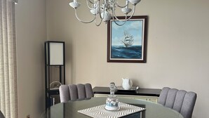 A dining area with a round glass table, grey upholstered chairs, a chandelier, and a framed seascape painting on the wall.
