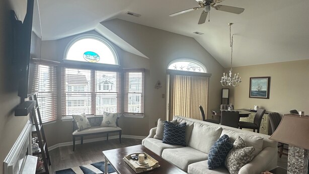 A spacious living room with a high ceiling, a ceiling fan, and a large window with blinds. There is a sofa, a coffee table, and a TV mounted on the wall.