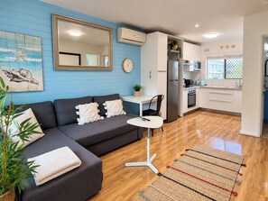 A modern living room with a grey sofa, a small white table, and a kitchen area with white cabinets and a stainless steel refrigerator.