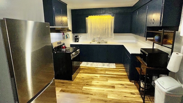 A compact kitchen with dark cabinets, a stainless steel refrigerator, a wooden floor, and a small sink area.
