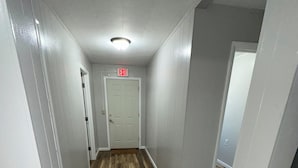 A hallway with wooden flooring, white walls, and a rug. There is an exit sign above a door.