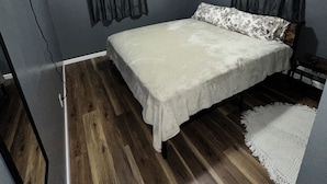 A bedroom with a bed, dark curtains, a ceiling light, and a wooden floor.