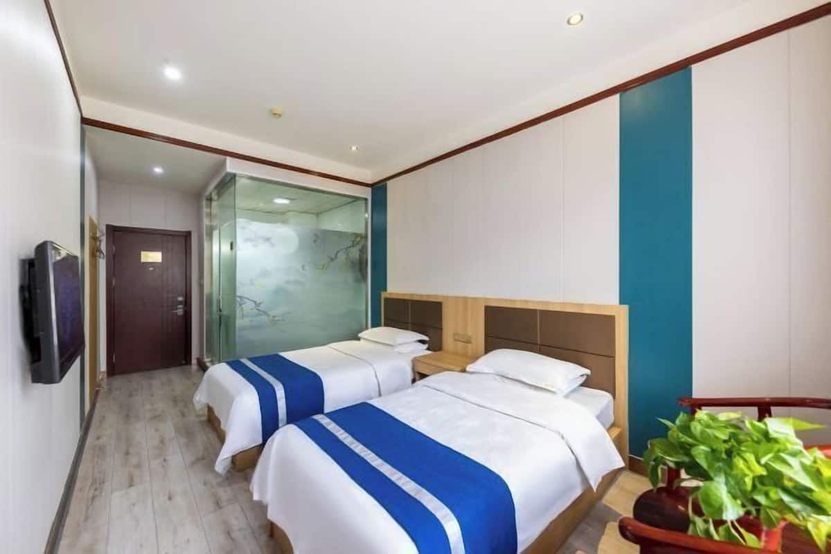 A hotel room with two beds, a TV, a glass shower enclosure, and a potted plant.