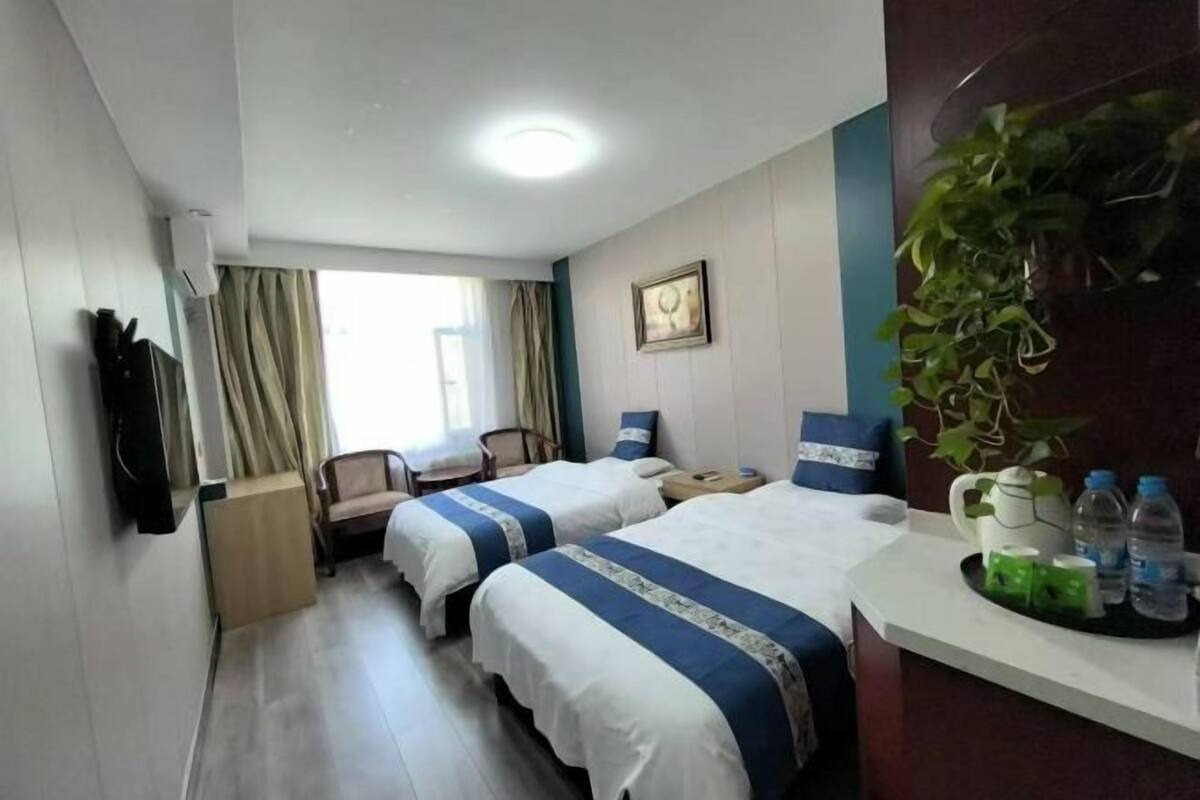 A hotel room with two beds, a desk, a chair, a plant, and a window with curtains.