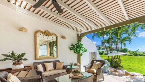 Un espace extérieur aménagé, abrité et doté de mobilier en osier, d’une table basse en verre et offrant une vue sur un paysage verdoyant luxuriant, parsemé de palmiers.