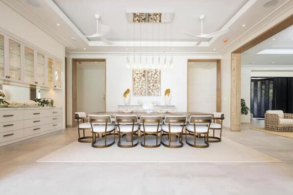 Un vaste espace dédié à la salle à manger, avec une longue table et six chaises, un lustre moderne et une grande fenêtre agrémentée de panneaux décoratifs.