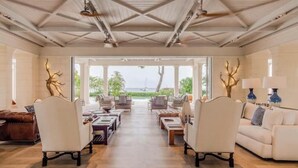 Un vaste salon offrant une vue sur l’océan, aménagé avec des canapés et des fauteuils blancs, et doté d’un plafond en bois.