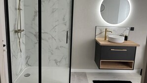 Et moderne badeværelse med et glasafskærmet brusebad, et rundt spejl med lys og en trækommode med en hvid vask.