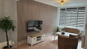 En moderne stue med et fladskærms-tv, en stor stueplante, fletmøbler og et vindue med skodder.