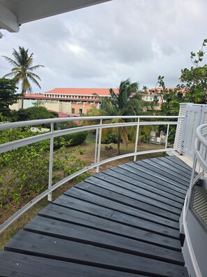 Um deck de madeira com guarda-corpo branco, com vista para um jardim ajardinado com palmeiras e um edifício com telhado vermelho.