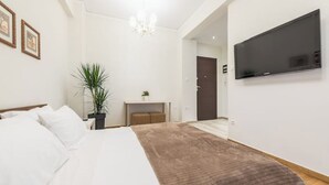 Une chambre moderne avec un grand lit, un téléviseur à écran plat fixé au mur, un petit bureau agrémenté d’une plante et une porte donnant sur une autre pièce.