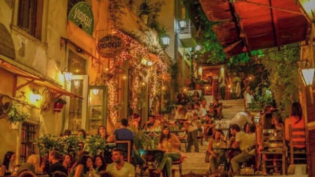 Un espace café en plein air, avec des tables et des chaises, entouré de bâtiments ornés d’enseignes et de verdure.